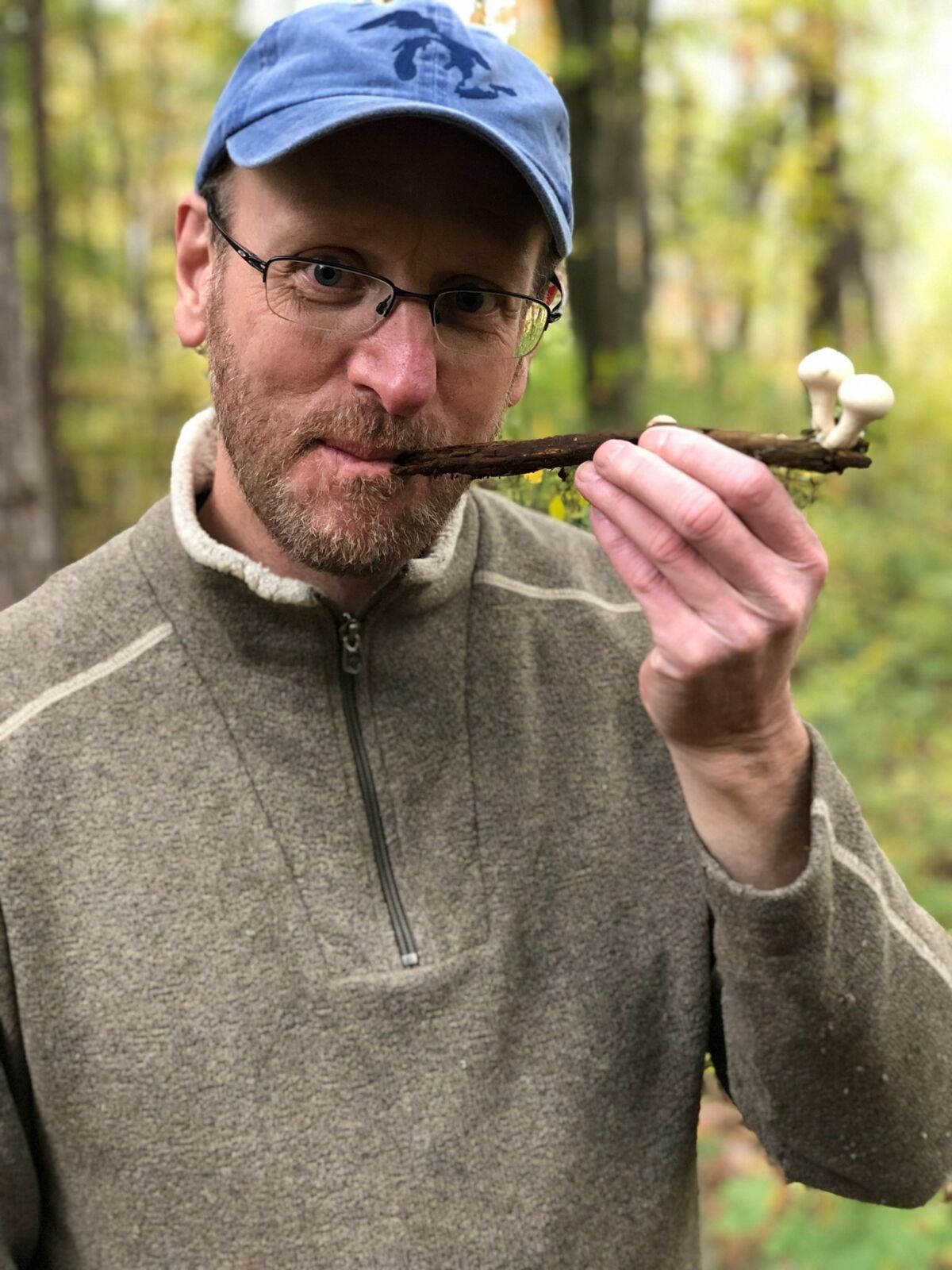 Andrew Lidral, wearing a hat and glasses, holds a stick with two mushrooms growing on the end like a pipe.