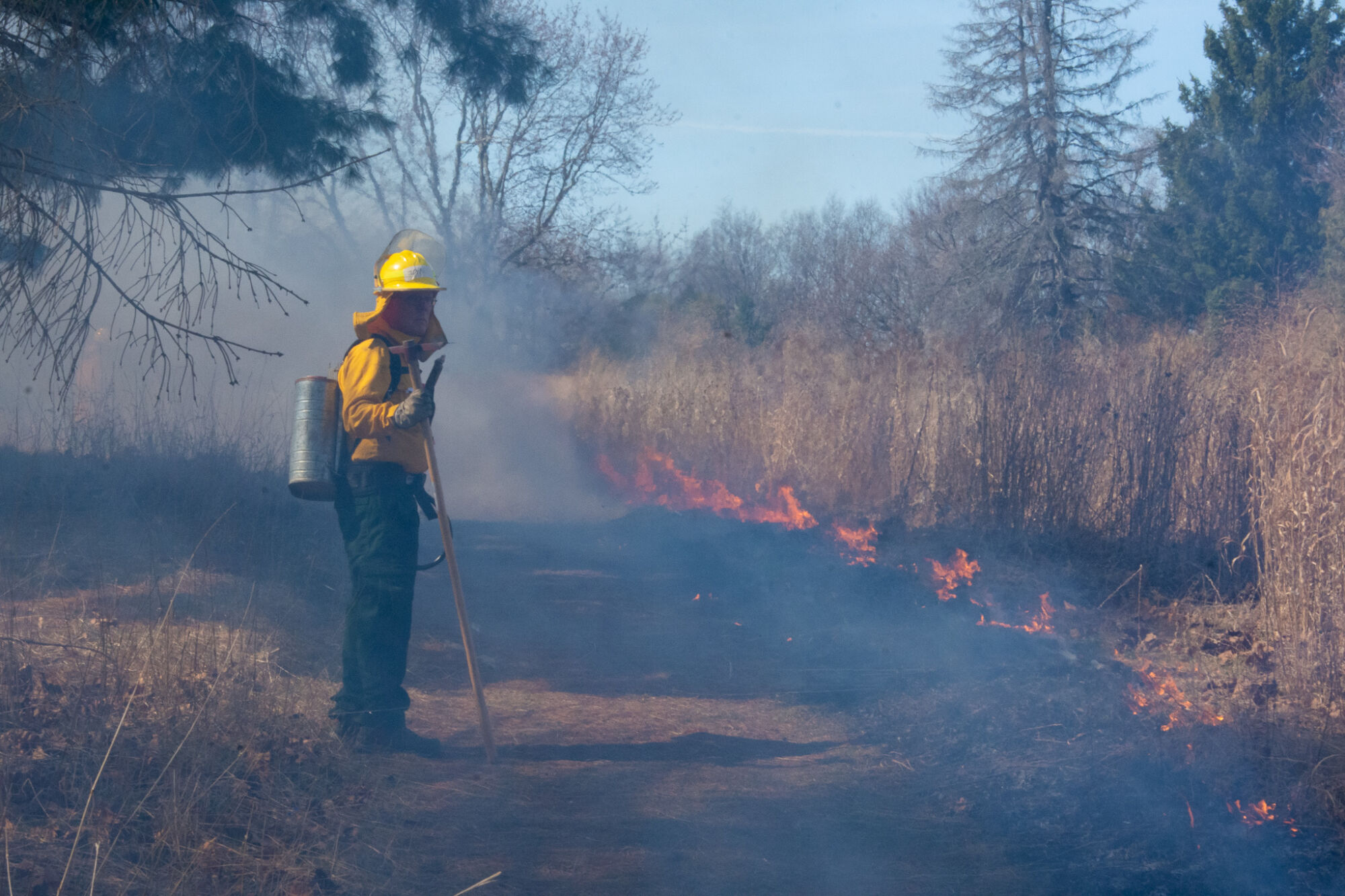 A prescribed burn crew volunteer stands by with a water backpack while prairie grasses burn at The Highlands.