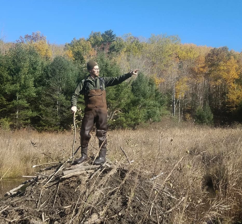 Photo of Chris Long standing in waders, looking and pointing to the right, standing before a forest in fall.